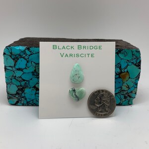 May include: Two pieces of green variscite gemstone, one teardrop shaped and one heart shaped, on a white card with the text "Black Bridge Variscite". A 25 pence coin is included for scale.
