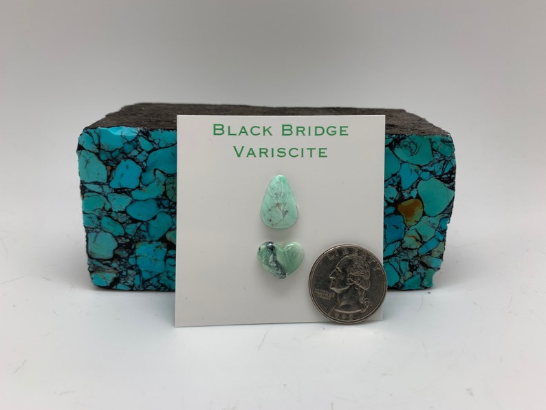 May include: Two pieces of green variscite gemstone, one teardrop shaped and one heart shaped, on a white card with the text "Black Bridge Variscite". A 25 pence coin is included for scale.