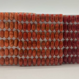 May include: Three rows of oval-shaped coral beads in shades of orange and red. The beads are arranged in a grid pattern on a white background.