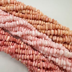May include: Close-up of several strands of coral beads in varying shades of pink and peach. The beads are small, irregular, and strung together, creating a textured surface. The beads are likely used for jewellery making.