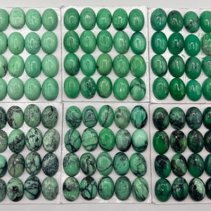 May include: A collection of 72 oval-shaped green gemstone cabochons, arranged in six rows of twelve. The cabochons are set in a white backing, and some have a black veining pattern.