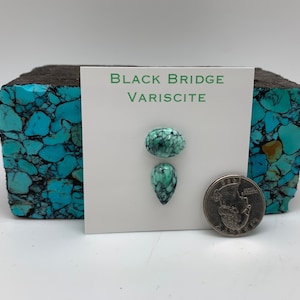 May include: Two green and black variscite cabochons, one oval and one teardrop shaped, are displayed on a white card with the text "BLACK BRIDGE VARISCITE". A US quarter is shown for scale.