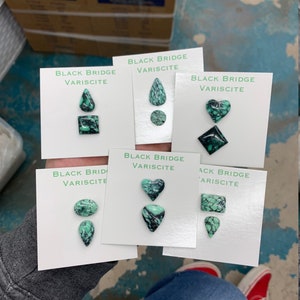 May include: Five white cards with the text "Black Bridge Variscite" and different green and black gemstone cabochons in the shapes of a teardrop, circle, heart, square, and rectangle.