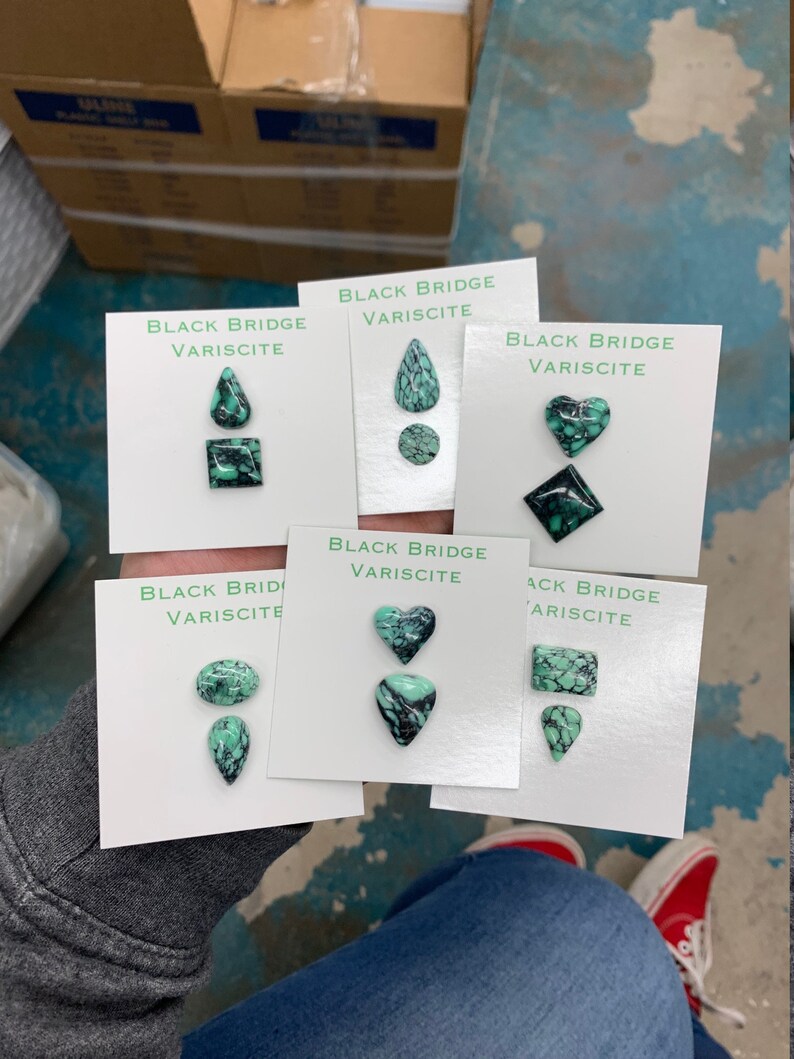 May include: Five white cards with the text "Black Bridge Variscite" and different green and black gemstone cabochons in the shapes of a teardrop, circle, heart, square, and rectangle.