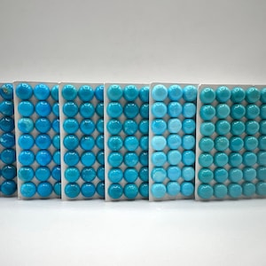 Puede incluir: Seis tarjetas rectangulares llenas de piedras preciosas redondas de turquesa pulida. Las piedras varían en tonos de azul, desde turquesa intenso hasta aguamarina claro. Las tarjetas están dispuestas en fila sobre un fondo blanco, mostrando las variaciones de color.