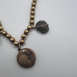 Puede incluir: Un collar de oro con un medall&oacute;n y un colgante de piedra gris. El medall&oacute;n tiene un dise&ntilde;o floral y un peque&ntilde;o c&iacute;rculo en el centro. El colgante de piedra tiene forma de coraz&oacute;n.