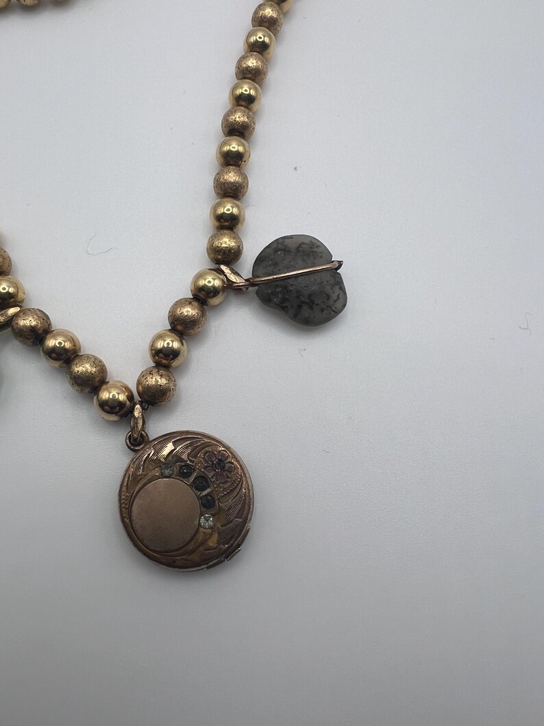 Puede incluir: Un collar de oro con un medall&oacute;n y un colgante de piedra gris. El medall&oacute;n tiene un dise&ntilde;o floral y un peque&ntilde;o c&iacute;rculo en el centro. El colgante de piedra tiene forma de coraz&oacute;n.