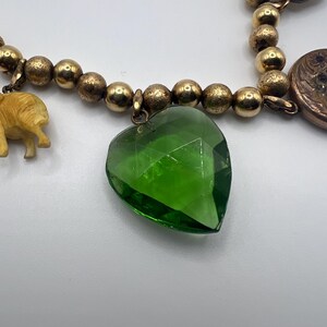 Puede incluir: Un collar de cadena dorada con m&uacute;ltiples dijes, incluyendo un dije en forma de coraz&oacute;n verde, una peque&ntilde;a figura de elefante, un dije redondo con forma de moneda y una piedra gris.