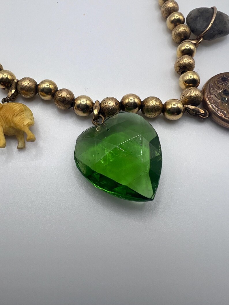 Puede incluir: Un collar de cadena dorada con m&uacute;ltiples dijes, incluyendo un dije en forma de coraz&oacute;n verde, una peque&ntilde;a figura de elefante, un dije redondo con forma de moneda y una piedra gris.