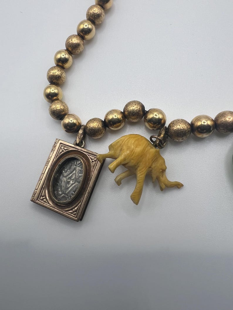 Puede incluir: Un collar de cuentas doradas con un peque&ntilde;o medall&oacute;n de oro y un dije de elefante de marfil tallado.