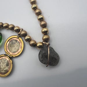 Puede incluir: Un collar de oro con una variedad de dijes, incluyendo un medall&oacute;n con una foto en su interior, una peque&ntilde;a cuenta de vidrio verde y una piedra gris envuelta en alambre.