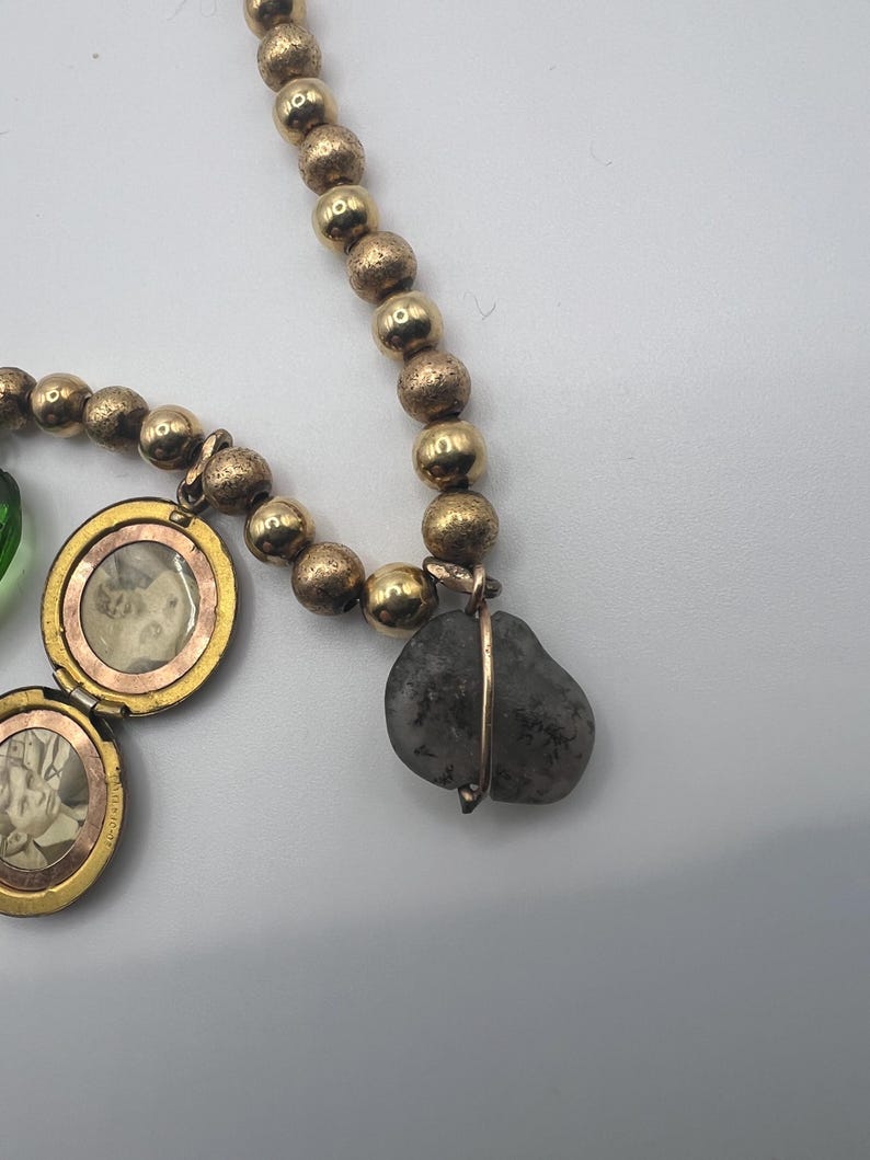 Puede incluir: Un collar de oro con una variedad de dijes, incluyendo un medall&oacute;n con una foto en su interior, una peque&ntilde;a cuenta de vidrio verde y una piedra gris envuelta en alambre.