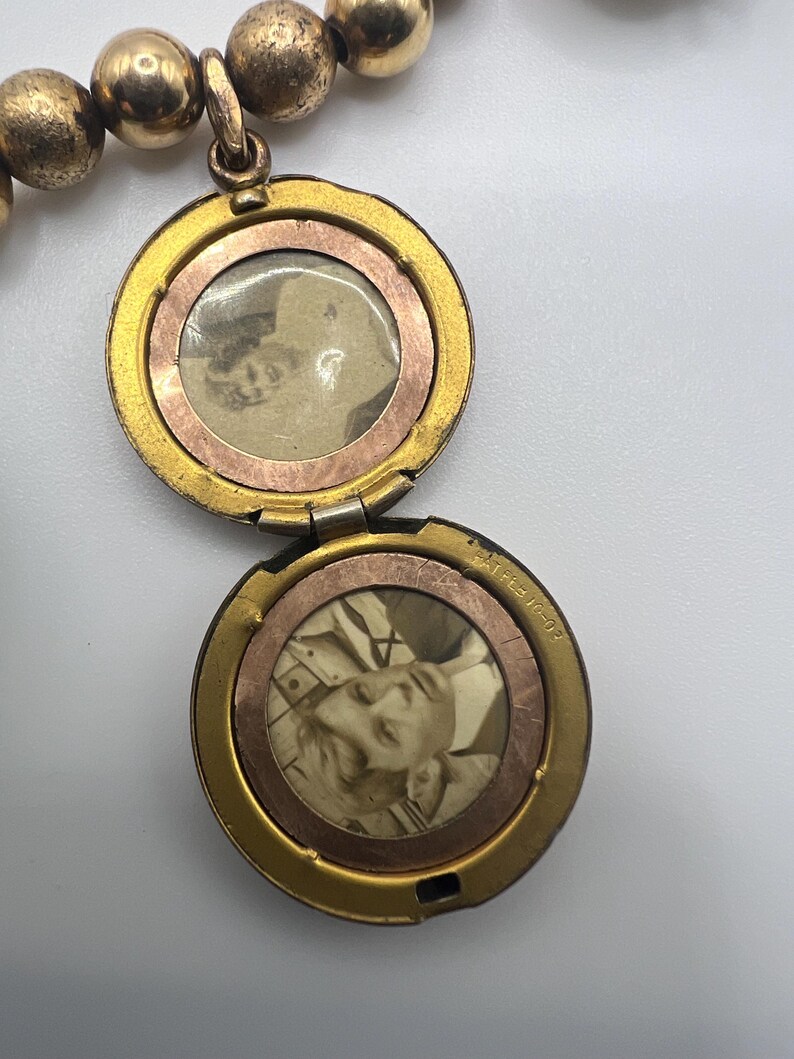 Puede incluir: Collar de oro con un medall&oacute;n redondo que se abre para revelar dos fotos. El medall&oacute;n est&aacute; unido a una cadena de cuentas doradas.