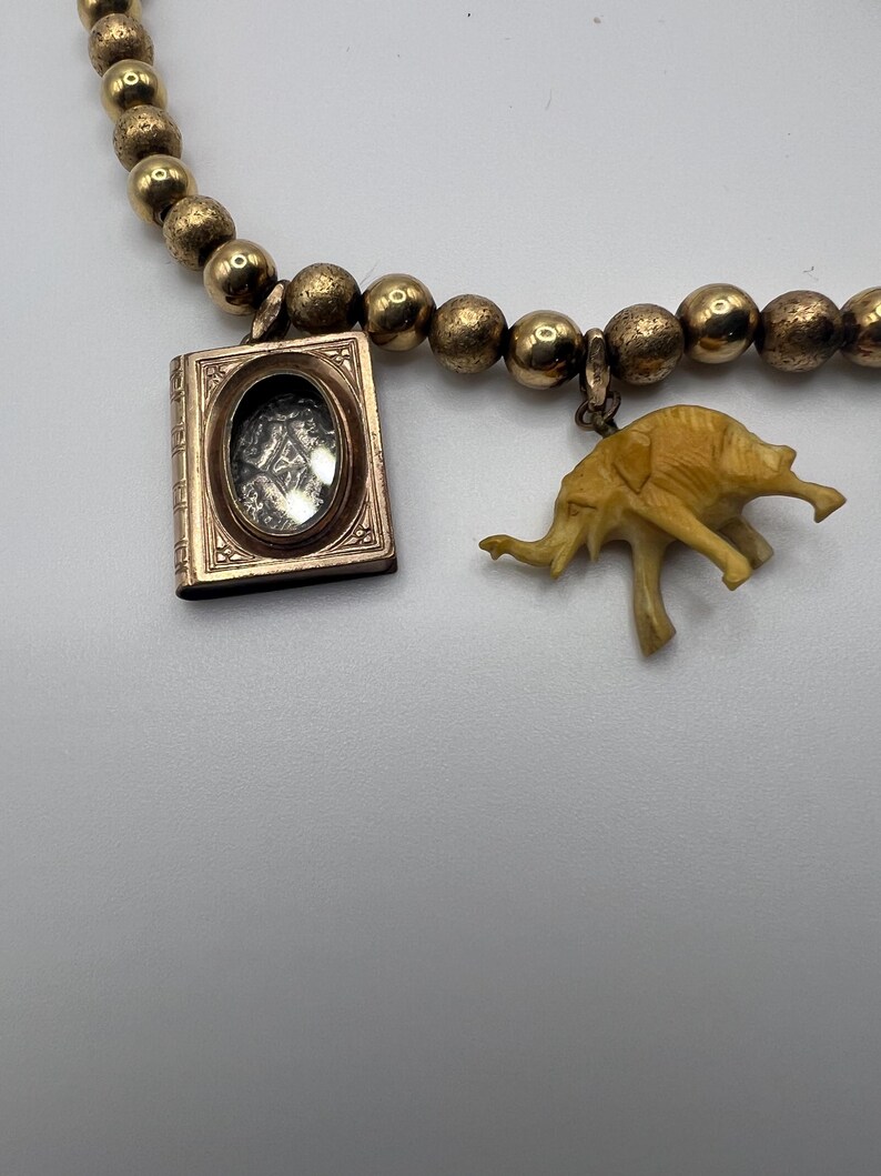 Puede incluir: Un collar de cadena de oro con un peque&ntilde;o medall&oacute;n y un dije de elefante de marfil tallado. El medall&oacute;n tiene una ventana de vidrio y un marco de oro.