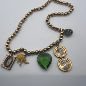 Puede incluir: Un collar de oro con una variedad de colgantes, incluyendo un cristal verde en forma de coraz&oacute;n, una peque&ntilde;a figura de elefante, un medall&oacute;n con una imagen en su interior y un peque&ntilde;o marco de oro con un dise&ntilde;o en su interior.