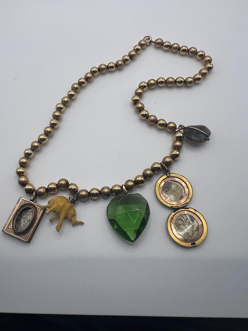 Puede incluir: Un collar de oro con una variedad de colgantes, incluyendo un cristal verde en forma de coraz&oacute;n, una peque&ntilde;a figura de elefante, un medall&oacute;n con una imagen en su interior y un peque&ntilde;o marco de oro con un dise&ntilde;o en su interior.