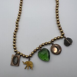 Puede incluir: Un collar de cuentas doradas con varios colgantes, incluyendo un colgante verde en forma de coraz&oacute;n, un peque&ntilde;o colgante de elefante, un medall&oacute;n con un dise&ntilde;o floral y un colgante de piedra gris.