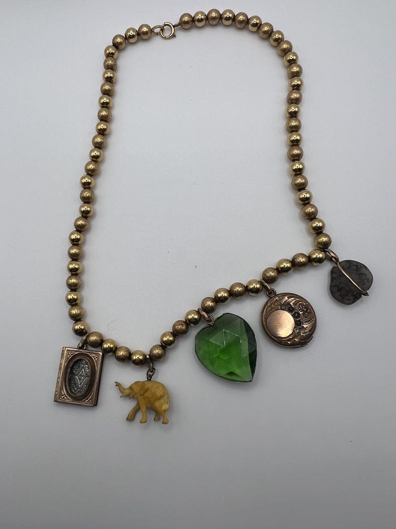 Puede incluir: Un collar de cuentas doradas con varios colgantes, incluyendo un colgante verde en forma de coraz&oacute;n, un peque&ntilde;o colgante de elefante, un medall&oacute;n con un dise&ntilde;o floral y un colgante de piedra gris.