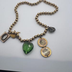 Puede incluir: Un collar de oro con m&uacute;ltiples encantos, incluyendo un colgante verde en forma de coraz&oacute;n, un peque&ntilde;o medall&oacute;n de oro, un peque&ntilde;o marco de imagen de oro, un peque&ntilde;o encanto de oro y un colgante de piedra gris.