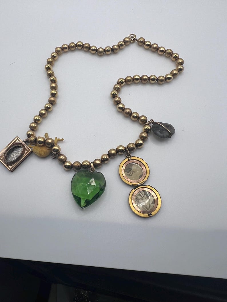 Puede incluir: Un collar de oro con m&uacute;ltiples encantos, incluyendo un colgante verde en forma de coraz&oacute;n, un peque&ntilde;o medall&oacute;n de oro, un peque&ntilde;o marco de imagen de oro, un peque&ntilde;o encanto de oro y un colgante de piedra gris.