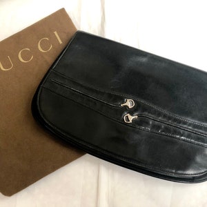 May include: A black leather Gucci clutch with silver hardware and a brown dust bag. The clutch has a curved shape and two silver clasps.
