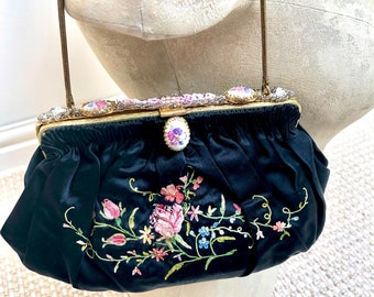 May include: Black satin clutch bag with embroidered flowers in pink, yellow, and blue. The clasp is decorated with rhinestones and oval floral details. A thin, gold-coloured chain strap is attached.