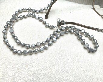 May include: A gray eyeglass chain featuring irregular, coin-shaped pearls interspersed with small, dark metallic beads. The chain attaches to the brown temples of a pair of glasses using clear silicone loops and silver-toned metal clasps.