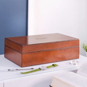 May include: A brown leather watch box with a hinged lid. The box is closed and sitting on a white surface.