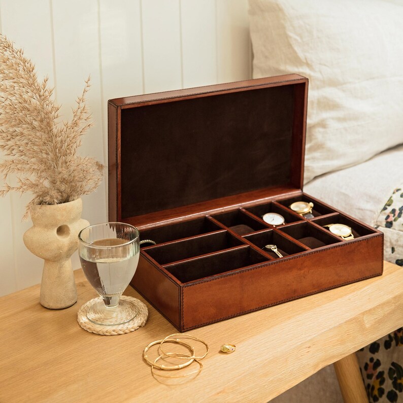 May include: A brown leather watch box with a hinged lid and multiple compartments for storing watches. The box is open and empty, revealing the compartments.