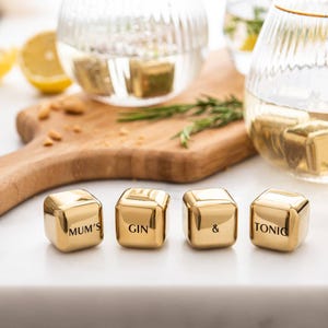 May include: Four gold-colored metal ice cubes with the words "MUM'S GIN & TONIC" engraved on them. The cubes are arranged in a row on a white surface. Two glasses with ice cubes are in the background, along with a wooden cutting board and a sprig of rosemary.