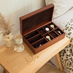 May include: A brown leather watch box with a lid open, revealing multiple compartments for storing watches and jewelry. The box is sitting on a wooden table with a glass of water and a ring on the surface.