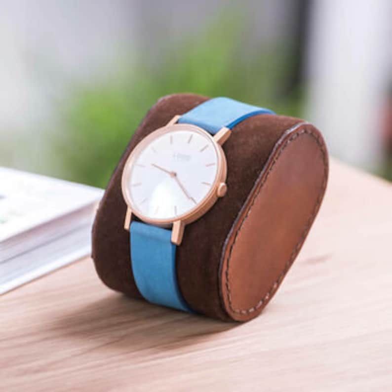 May include: A wristwatch with a white face and rose gold hands and case, on a blue leather strap, displayed on a brown leather watch stand.