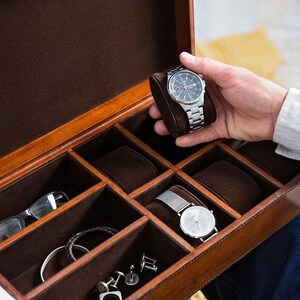 May include: A brown leather watch box with multiple compartments for storing watches, cufflinks, and other accessories. The box is open and a silver watch is being placed in one of the compartments.