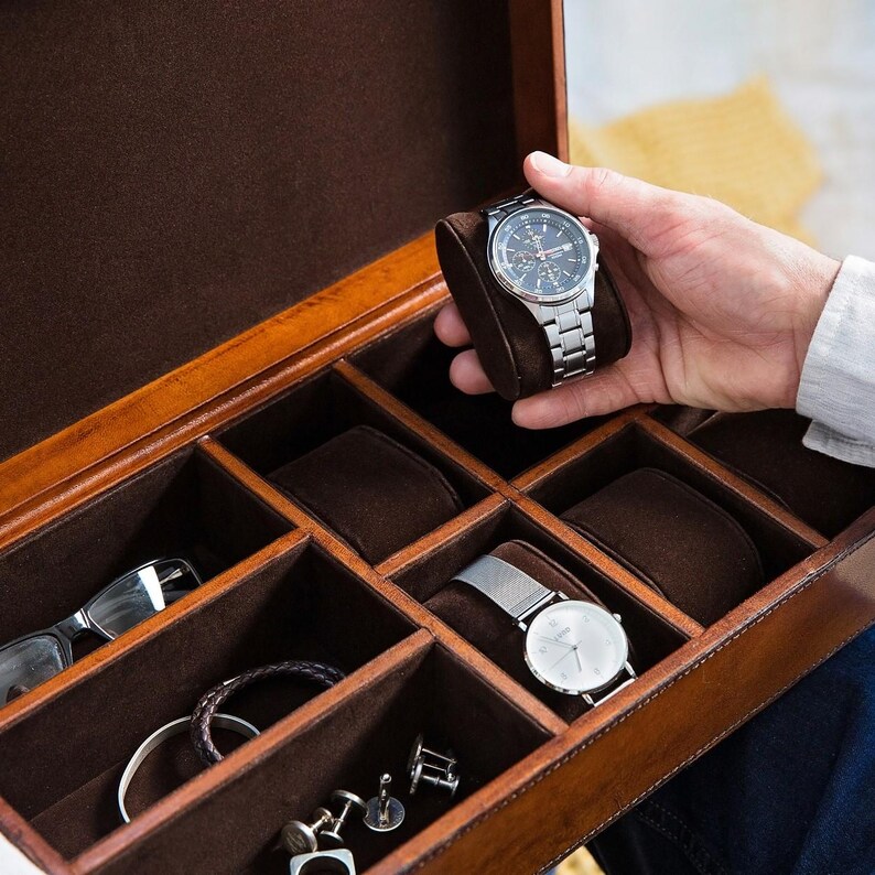 May include: A brown leather watch box with multiple compartments for storing watches, cufflinks, and other accessories. The box is open and a silver watch is being placed in one of the compartments.