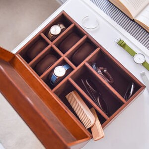 May include: A wooden watch box with a hinged lid, featuring eight compartments lined with brown fabric. The box is open, revealing a comb and sunglasses in two of the compartments. There are two watches in the compartments.