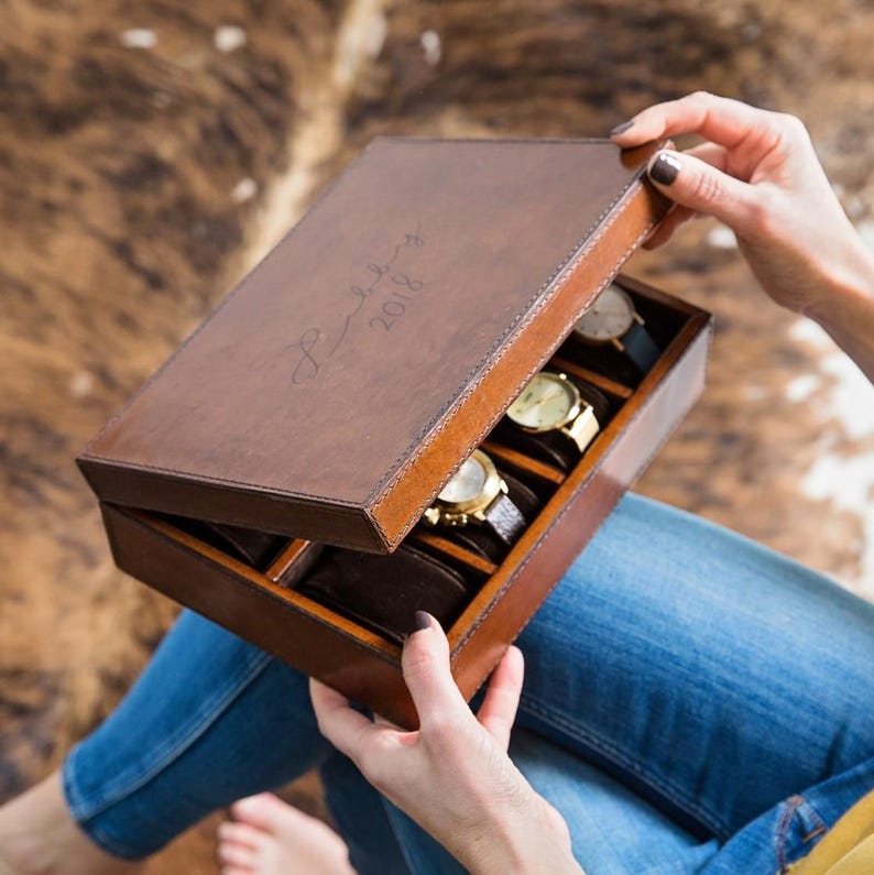 May include: A brown leather watch box with a personalized inscription "Established 2018". The box is open and contains several watches with gold bands.