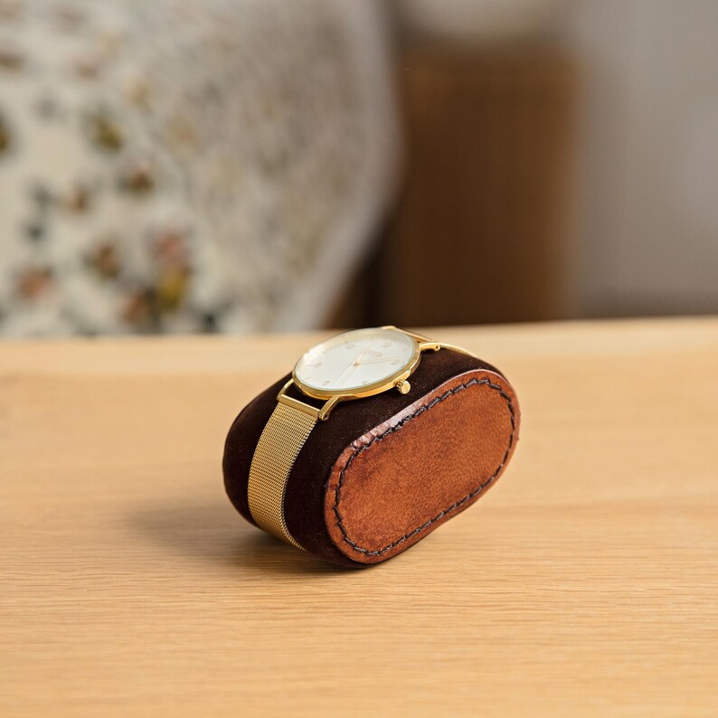 May include: A gold watch with a white face and a mesh band is displayed on a brown leather watch box.