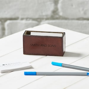 May include: A brown leather business card holder with the text "SMITH AND SONS" on the front. The holder is rectangular and sits on a white surface. Two blue pens and a stack of business cards are also visible.