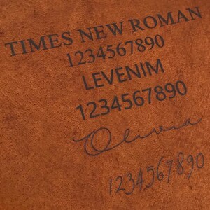 May include: Close-up of a brown leather journal with the text "TIMES NEW ROMAN" and "LEVENIM" embossed on the cover. The numbers "1234567890" are also embossed on the cover.