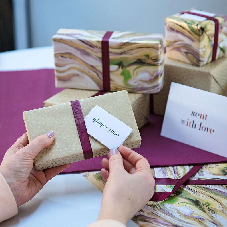 May include: A gift wrapped in brown paper with gold flecks and a burgundy ribbon. The gift is topped with a white card that reads "ginger rose". A card with the text "sent with love" is visible in the background.