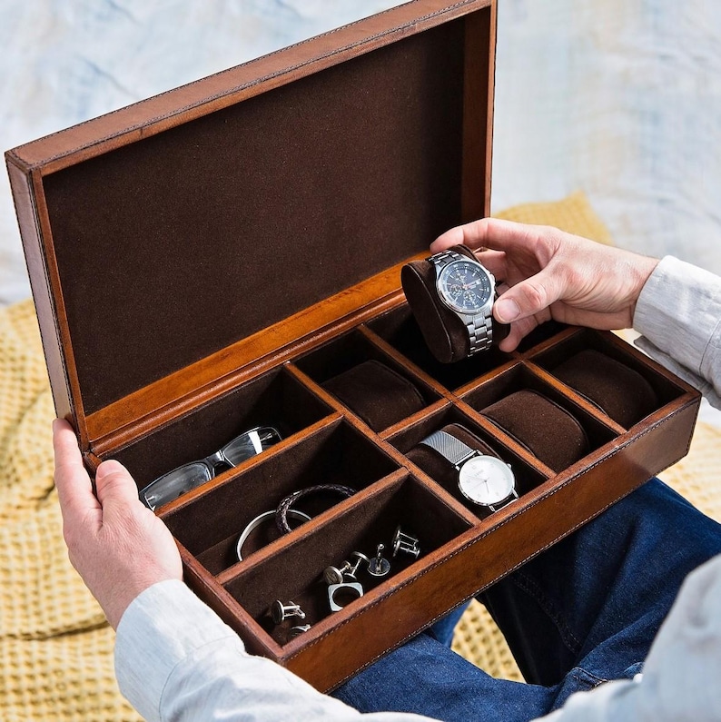 May include: A brown leather watch box with multiple compartments for storing watches, cufflinks, and other accessories. The box is open and a silver watch is being placed inside.