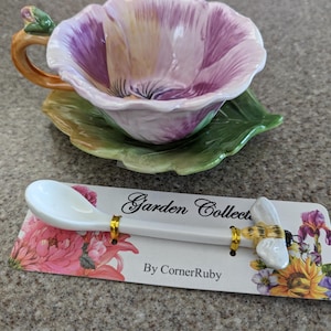 May include: A floral-shaped teacup and saucer set with a matching spoon. The teacup is purple and white, resembling a flower, with a green leaf-shaped saucer. The spoon has a bee design and the text "Garden Collection By CornerRuby".
