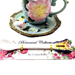Puede incluir: Juego de taza y platillo decorativo con diseño floral y de pájaros. La taza es azul claro con un acento floral rosa y amarillo. El platillo tiene un borde dorado y una pequeña figura de pájaro. También se incluye una cuchara con un acento de mariposa.