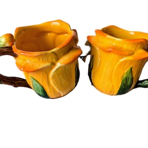 Könnte beinhalten: Eine weiße Keramik-Tasse mit einem farbenfrohen Blumenmuster. Die Tasse zeigt eine lebendige Anordnung von rosa, lila und gelben Blumen auf weißem Hintergrund. Sie hat einen Standardgriff und eine glatte, glänzende Oberfläche.