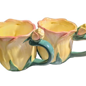 May include: Two ceramic mugs shaped like yellow tulip flowers with green handles and accents. The rims are pink, and the interior is yellow. Each mug has a small flower bud detail on the handle.