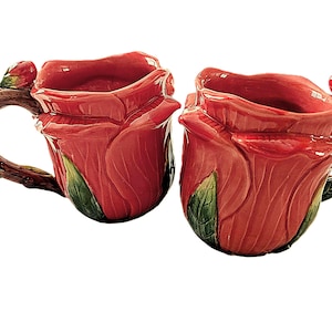 May include: Two ceramic mugs shaped like red roses. The mugs have a detailed design with green leaves and brown branch-like handles. The mugs are set against a white background.