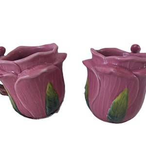 May include: A pink ceramic teacup and sugar bowl set, each shaped like a rose. The teacup has a brown handle and green leaves are painted on the side. The sugar bowl has a small knob on the lid.