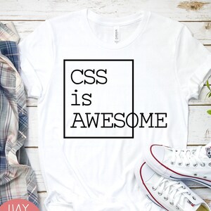 May include: A white short-sleeved t-shirt featuring a black square outline containing the text CSS is AWESOME in a clean sans-serif font. The shirt is displayed flat with a plaid flannel shirt and white canvas trainers visible nearby.