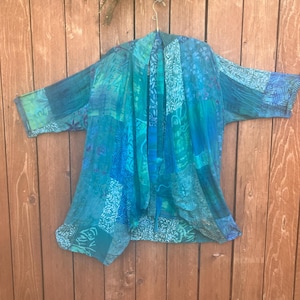May include: A lightweight open-front kimono jacket featuring a patchwork design in various shades of blue, teal, and green. The garment displays a mix of botanical and abstract patterns across its fabric panels, with three-quarter length sleeves.