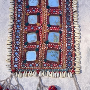 May include: A rectangular textile wall hanging featuring a red fabric base with six square mirrors arranged in two columns. The border is decorated with white cowrie shells, small circular mirrors, and colorful thread accents. Three beaded tassels with multi-colored yarn pom-poms hang from the bottom edge.